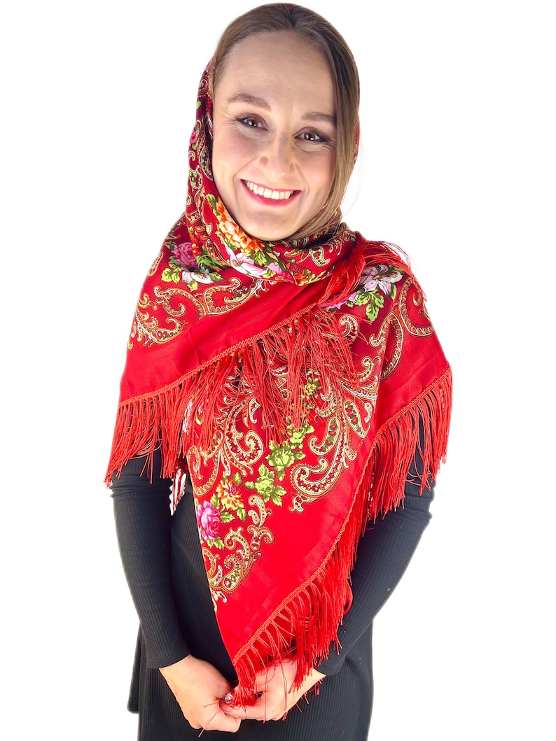 May include: A red headscarf with a floral pattern and fringe. The scarf is worn by a person with brown hair and a black shirt.
