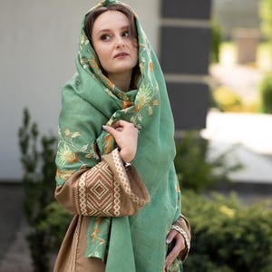 May include: A woman wearing a green shawl with floral embroidery. The shawl is draped over her head and shoulders, and she is looking off to the side. She is wearing a brown dress with embroidered details.