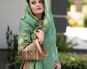 May include: A woman wearing a green shawl with floral embroidery. The shawl is draped over her head and shoulders, and she is looking off to the side. She is wearing a brown dress with embroidered details.