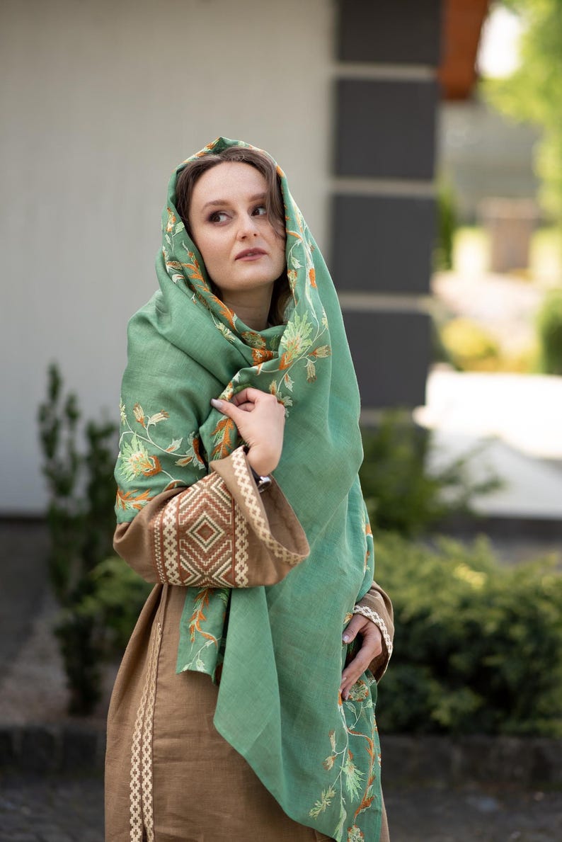 May include: A woman wearing a green shawl with floral embroidery. The shawl is draped over her head and shoulders, and she is looking off to the side. She is wearing a brown dress with embroidered details.