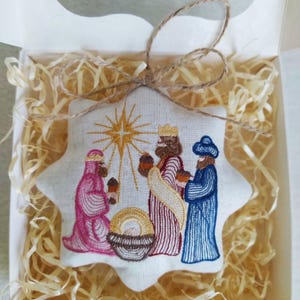 May include: Embroidered star-shaped Christmas ornament featuring the three wise men, a star, and a manger. The ornament is in a white box with straw-like filler, tied with twine. The figures are in shades of pink, blue, and brown.