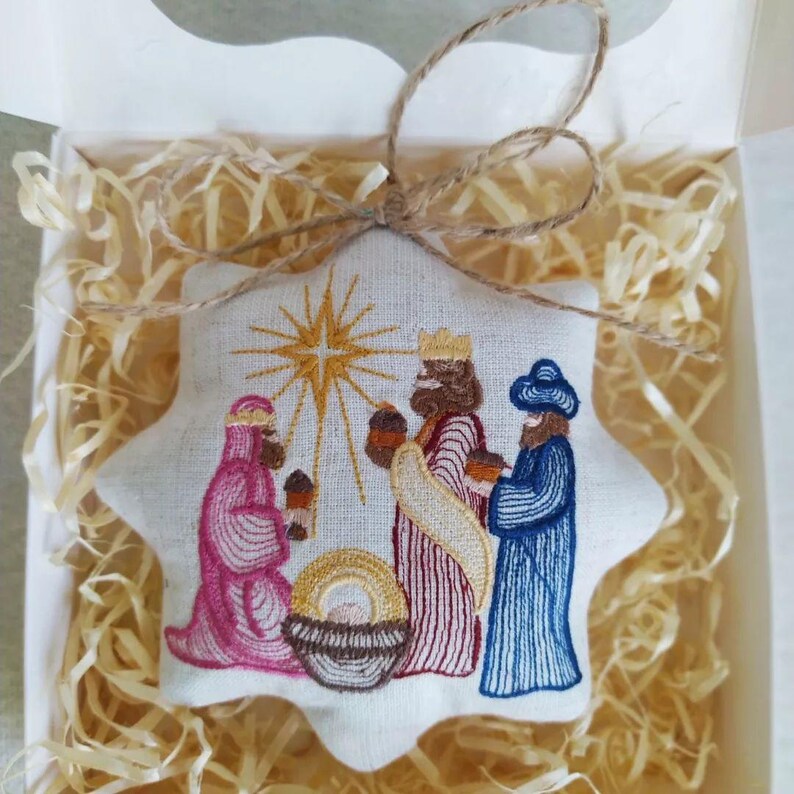 May include: Embroidered star-shaped Christmas ornament featuring the three wise men, a star, and a manger. The ornament is in a white box with straw-like filler, tied with twine. The figures are in shades of pink, blue, and brown.