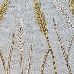 Op de afbeelding: Close-up van geborduurde tarwestengels in goud en gebroken wit garen op een lichtgrijze linnen stof. Het tarweontwerp is gedetailleerd, met elke stengel en aar zorgvuldig gestikt, waardoor een textuurrijk en elegant patroon ontstaat.