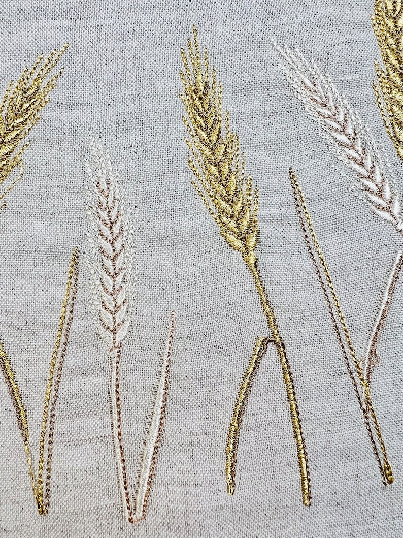 Op de afbeelding: Close-up van geborduurde tarwestengels in goud en gebroken wit garen op een lichtgrijze linnen stof. Het tarweontwerp is gedetailleerd, met elke stengel en aar zorgvuldig gestikt, waardoor een textuurrijk en elegant patroon ontstaat.