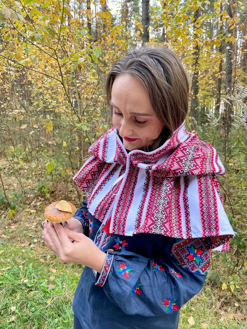 May include: A woman wearing a traditional embroidered cape and blouse, holding a mushroom with a leaf in a forest setting. The cape features a red and white geometric pattern. The blouse has colorful floral embroidery.