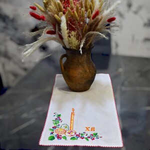 Puede incluir: Un camino de mesa blanco bordado con un borde rojo. El bordado presenta una vela, tres huevos y las letras cir&iacute;licas "XB". Un jarr&oacute;n con flores secas se encuentra sobre el camino de mesa.
