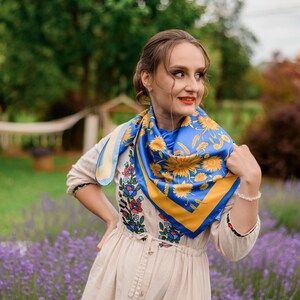 May include: A woman wearing a beige dress with embroidered flowers and a blue and yellow floral patterned scarf. She is standing in a field of lavender.