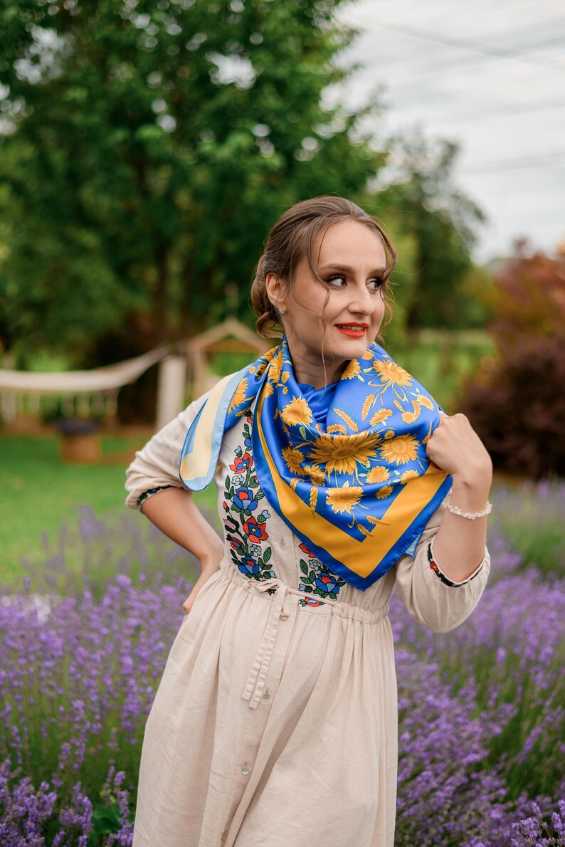 May include: A woman wearing a beige dress with embroidered flowers and a blue and yellow floral patterned scarf. She is standing in a field of lavender.