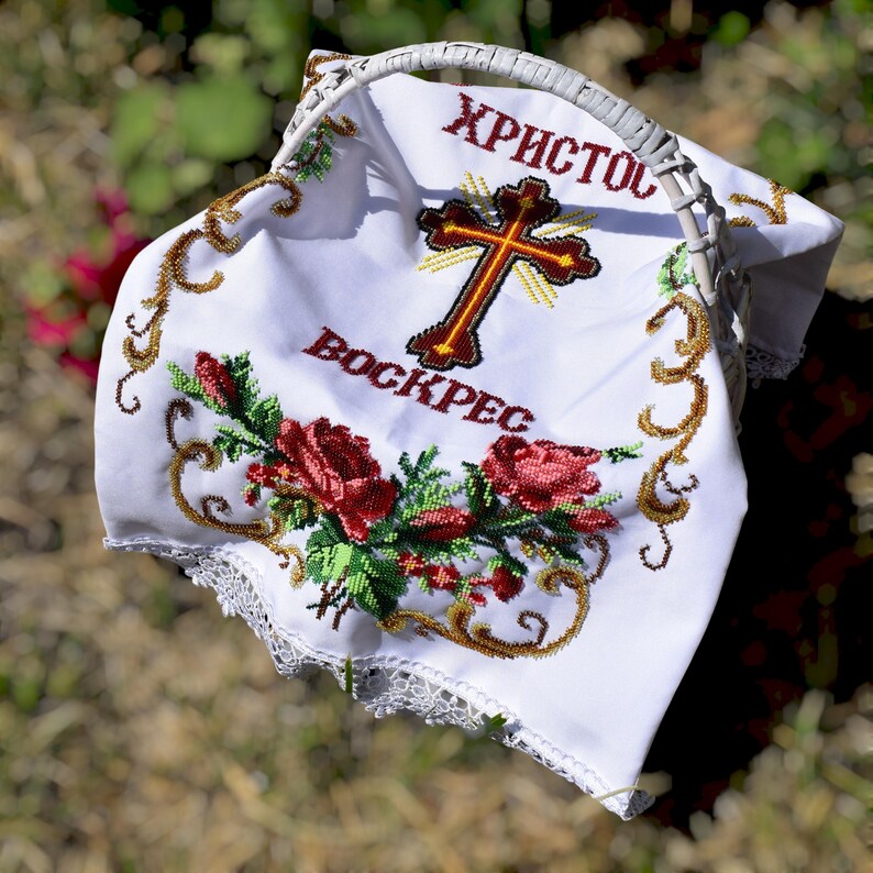 K&ouml;nnte beinhalten: Ein wei&szlig;er Osterkorb mit Henkel, bedeckt mit einem wei&szlig;en bestickten Tuch. Das Tuch zeigt ein Kreuz, die Worte "Christus ist auferstanden" in Rot und ein Blumendesign mit roten Rosen und gr&uuml;nen Bl&auml;ttern. Der Korb steht auf einer grasbewachsenen Oberfl&auml;che.