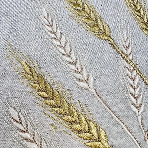 Op de afbeelding: Close-up van geborduurde tarwestengels in goud en gebroken wit op een lichtgrijze stof. Het tarweontwerp is gedetailleerd, met elke stengel schuin over het frame, waardoor een textuurrijk en elegant patroon ontstaat.
