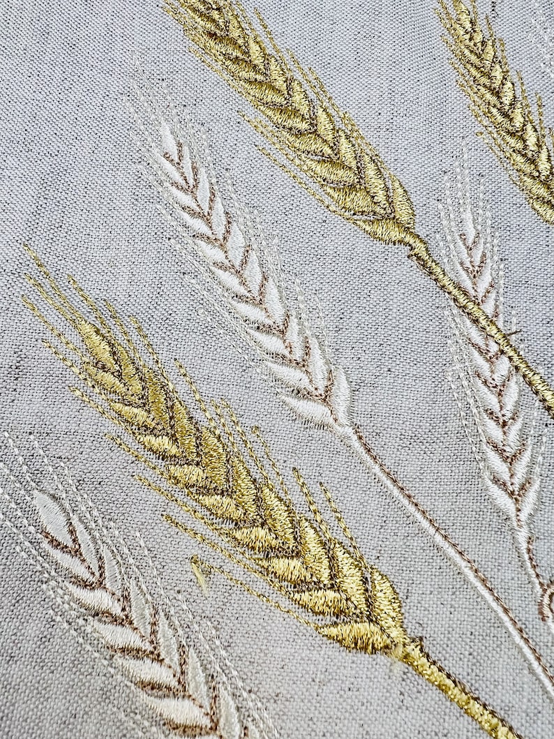 Op de afbeelding: Close-up van geborduurde tarwestengels in goud en gebroken wit op een lichtgrijze stof. Het tarweontwerp is gedetailleerd, met elke stengel schuin over het frame, waardoor een textuurrijk en elegant patroon ontstaat.