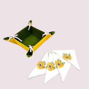 May include: A set of four white linen napkins with embroidered sunflower designs. The napkins are folded in a triangular shape and are arranged on a green and yellow felt basket.