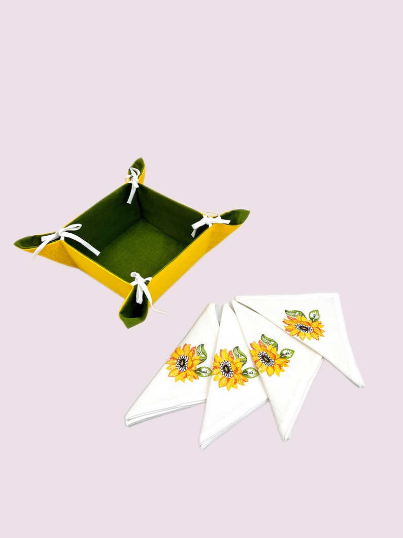 May include: A set of four white linen napkins with embroidered sunflower designs. The napkins are folded in a triangular shape and are arranged on a green and yellow felt basket.