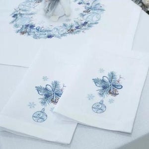 K&ouml;nnte beinhalten: Wei&szlig;e Tischw&auml;sche und Servietten zum Thema Weihnachten. Die Tischdecke zeigt ein blau-wei&szlig;es Kranzdesign. Zwei wei&szlig;e Servietten sind mit einer blauen Schleife, Tannennadeln und einer Weihnachtskugel verziert.