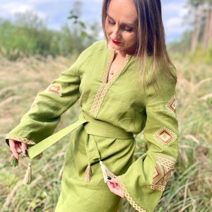 May include: A long, green linen dress with embroidered details in gold and brown. The dress has a belt with tassels and long sleeves.