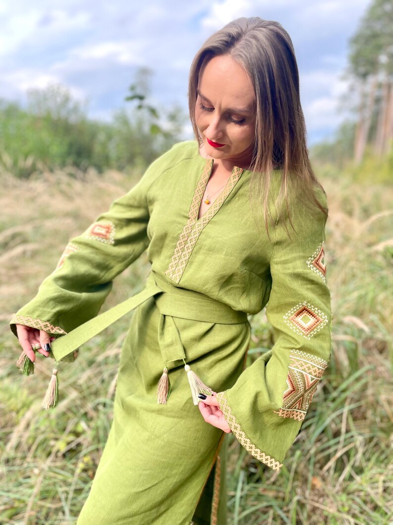 May include: A long, green linen dress with embroidered details in gold and brown. The dress has a belt with tassels and long sleeves.