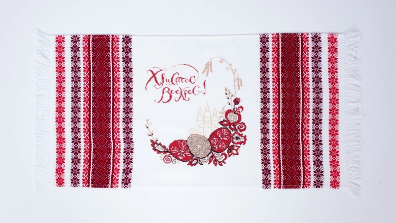 Puede incluir: Camino de mesa bordado blanco de Pascua con patrones florales y geom&eacute;tricos rojos y burdeos. El centro presenta una corona con huevos de Pascua y texto en escritura roja. Los bordes tienen flecos.