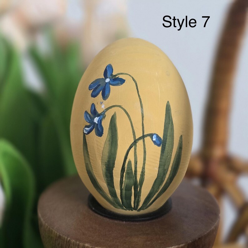 May include: A hand-painted, egg-shaped decorative item. The egg is painted yellow with blue floral accents and green leaves. The text "Style 7" is displayed in black. The egg is mounted on a wooden base.