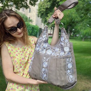 K&ouml;nnte beinhalten: Eine taupefarbene Tragetasche mit einem floralen Stickdesign in Wei&szlig; und Hellblau. Die Tasche hat einen breiten Schulterriemen und eine plissierte Vorderseite. Der Hintergrund ist eine gr&uuml;ne, &auml;u&szlig;ere Umgebung.