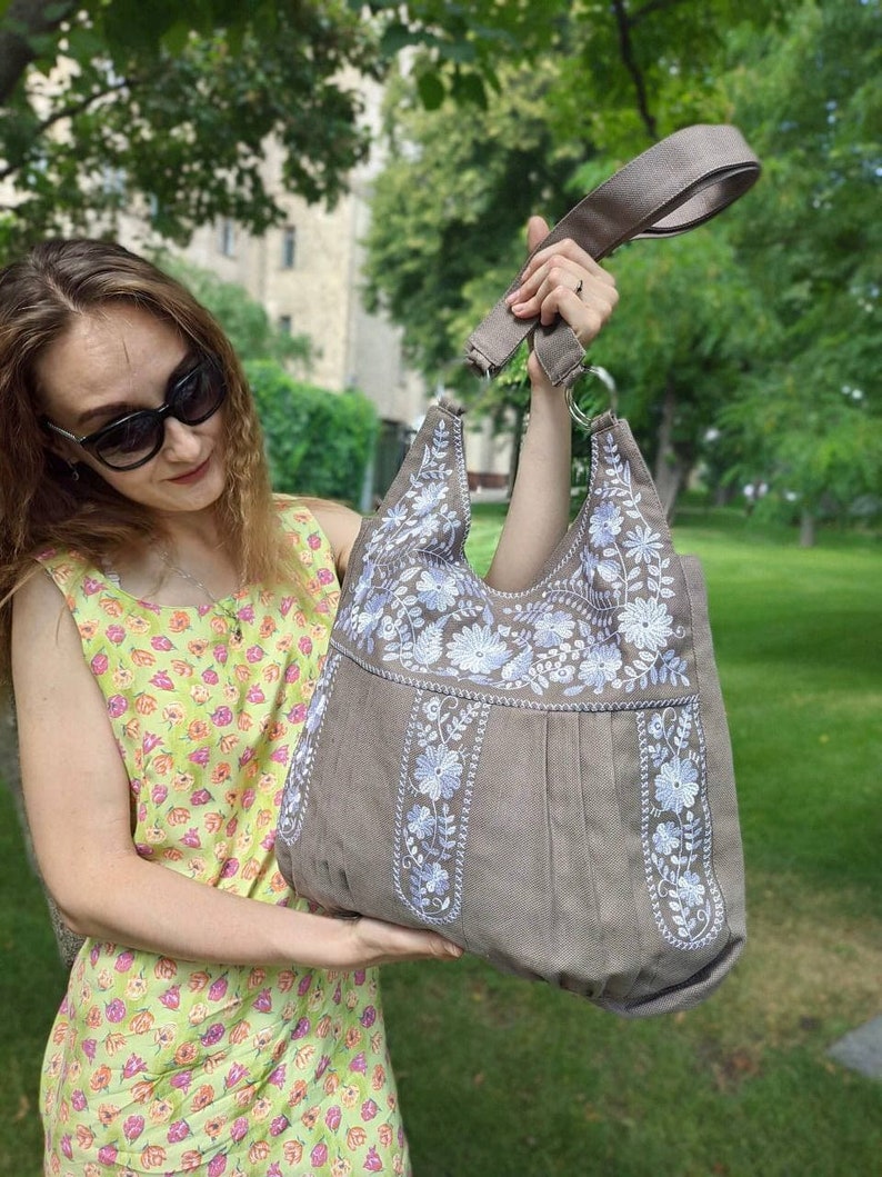 K&ouml;nnte beinhalten: Eine taupefarbene Tragetasche mit einem floralen Stickdesign in Wei&szlig; und Hellblau. Die Tasche hat einen breiten Schulterriemen und eine plissierte Vorderseite. Der Hintergrund ist eine gr&uuml;ne, &auml;u&szlig;ere Umgebung.