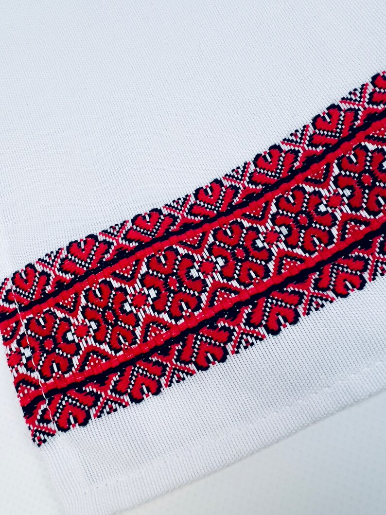 May include: White fabric with a red and black embroidered border. The embroidery features a geometric pattern.