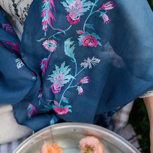 May include: A teal blue scarf with pink and blue floral embroidery. The scarf is draped over a white surface and a silver bowl filled with water and peach-coloured roses.