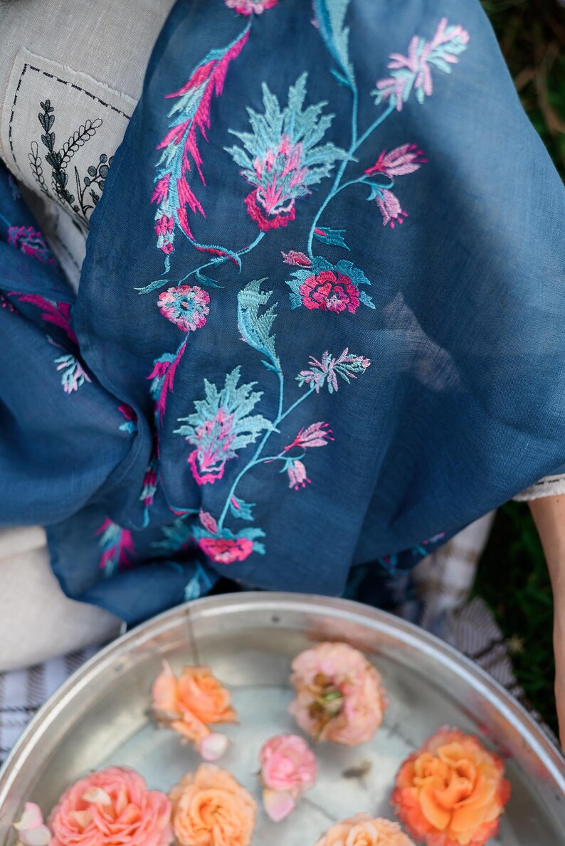 May include: A teal blue scarf with pink and blue floral embroidery. The scarf is draped over a white surface and a silver bowl filled with water and peach-coloured roses.