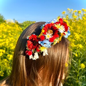Pode incluir: Uma faixa de cabe&ccedil;a floral com flores vermelhas, amarelas e azuis. A faixa de cabe&ccedil;a &eacute; usada por uma pessoa com cabelo castanho comprido. O fundo &eacute; um campo de flores amarelas.