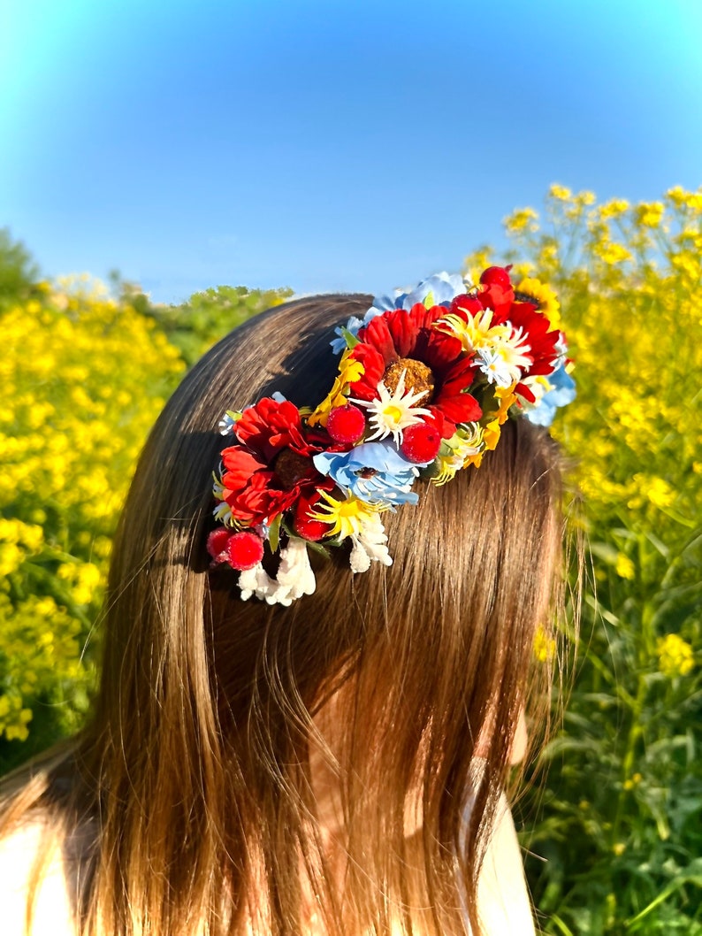 Pode incluir: Uma faixa de cabe&ccedil;a floral com flores vermelhas, amarelas e azuis. A faixa de cabe&ccedil;a &eacute; usada por uma pessoa com cabelo castanho comprido. O fundo &eacute; um campo de flores amarelas.
