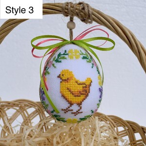 Op de afbeelding: Paasei-ornament met een kruissteekontwerp van een gele kuiken, omgeven door bloemenborduurwerk. Het ei wordt opgehangen door een groen en roze lint en een lus van jute touw. De tekst "Style 3" staat in de linkerbovenhoek.