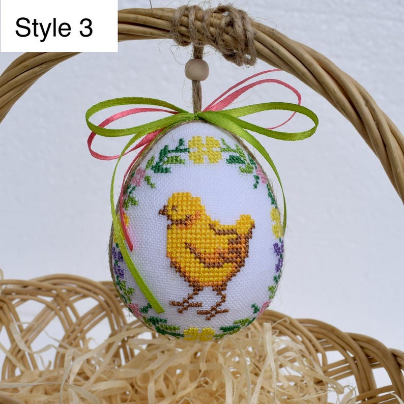 Op de afbeelding: Paasei-ornament met een kruissteekontwerp van een gele kuiken, omgeven door bloemenborduurwerk. Het ei wordt opgehangen door een groen en roze lint en een lus van jute touw. De tekst "Style 3" staat in de linkerbovenhoek.