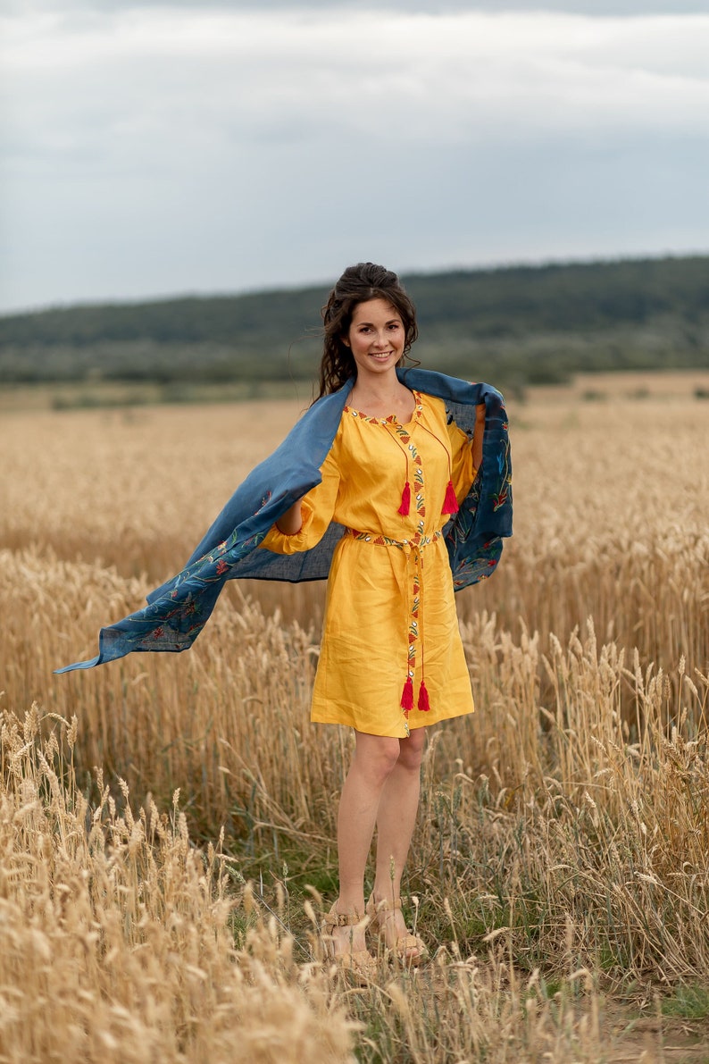 May include: A woman in a yellow dress with embroidered details and a blue scarf stands in a field of wheat. She is smiling and looking at the camera.