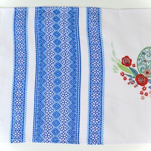 May include: White rectangular table runner with blue geometric patterned borders and fringe. The runner features embroidered Easter eggs and floral designs in green, red, and yellow.