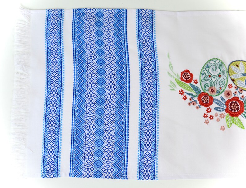 May include: White rectangular table runner with blue geometric patterned borders and fringe. The runner features embroidered Easter eggs and floral designs in green, red, and yellow.