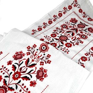 K&ouml;nnte beinhalten: Wei&szlig;er Tischl&auml;ufer aus Leinen mit roten und schwarzen gestickten Blumendesigns. Das Design zeigt ein sich wiederholendes Muster aus stilisierten Blumen und Bl&auml;ttern.