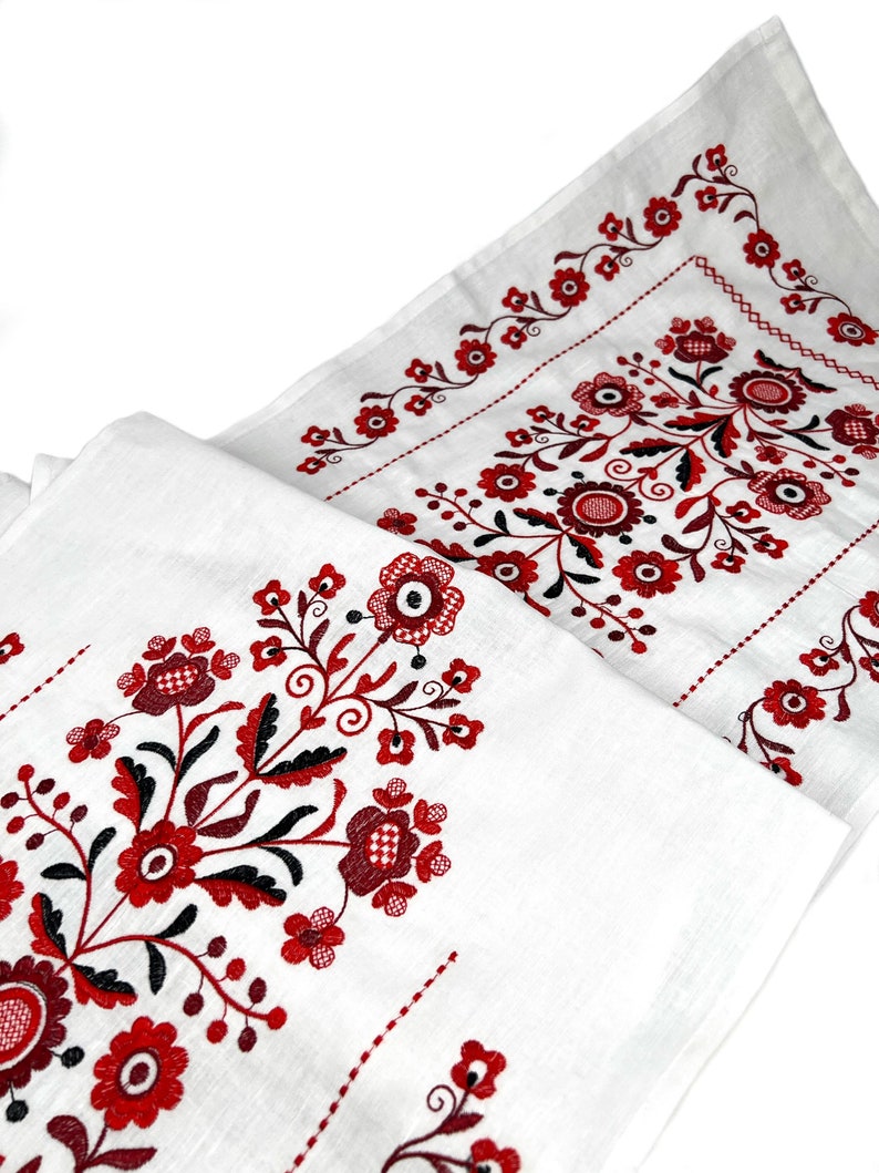 K&ouml;nnte beinhalten: Wei&szlig;er Tischl&auml;ufer aus Leinen mit roten und schwarzen gestickten Blumendesigns. Das Design zeigt ein sich wiederholendes Muster aus stilisierten Blumen und Bl&auml;ttern.