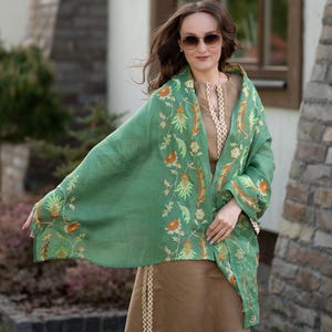 May include: A woman wearing a green shawl with floral embroidery and a brown skirt. The shawl has a light green background with orange and yellow flowers.