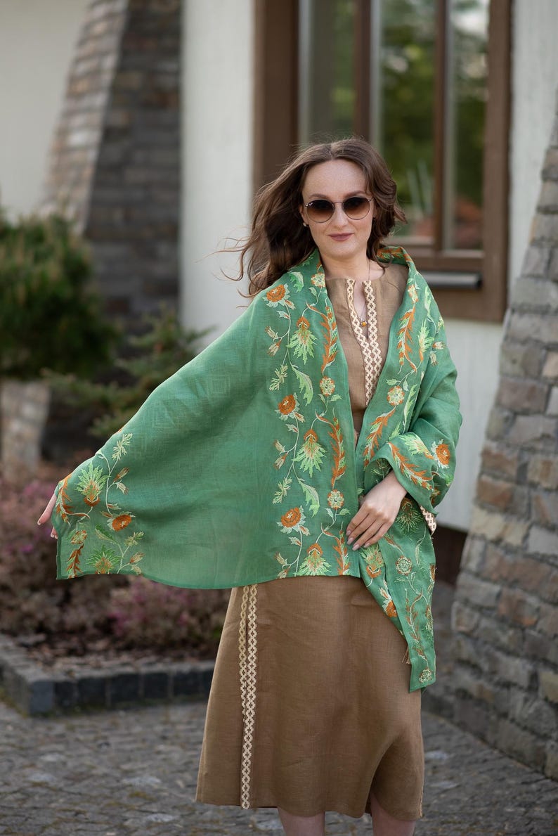 May include: A woman wearing a green shawl with floral embroidery and a brown skirt. The shawl has a light green background with orange and yellow flowers.