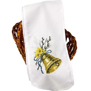 K&ouml;nnte beinhalten: Wei&szlig;es besticktes K&uuml;chentuch mit einer goldenen Glocke, einem blauen Band und gelben Blumen. Das Tuch ist gefaltet und in einem braunen Weidenkorb gelegt.