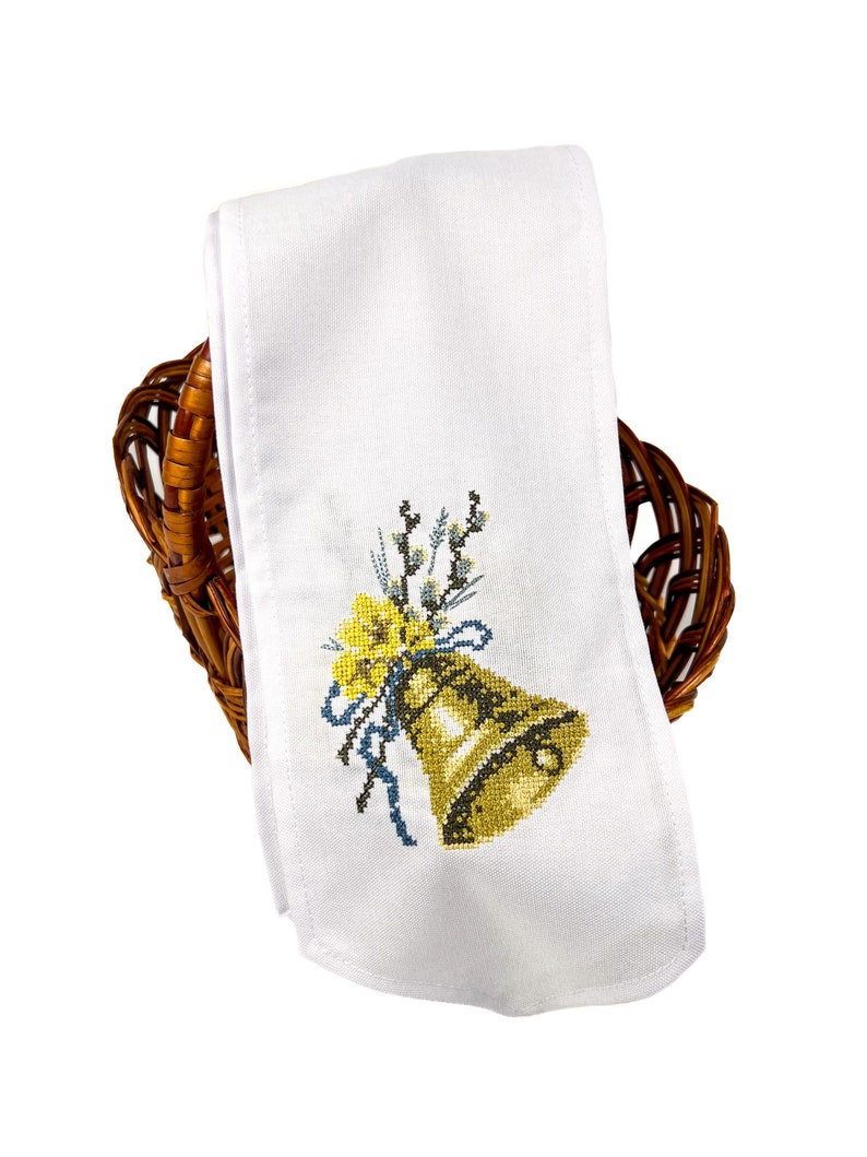 K&ouml;nnte beinhalten: Wei&szlig;es besticktes K&uuml;chentuch mit einer goldenen Glocke, einem blauen Band und gelben Blumen. Das Tuch ist gefaltet und in einem braunen Weidenkorb gelegt.