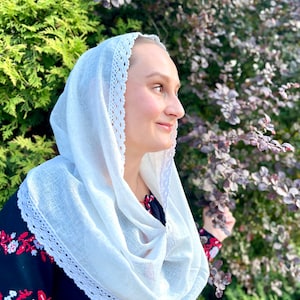May include: A white headscarf with a delicate lace trim, draped around the neck. The scarf is paired with a black top featuring red and white embroidered floral patterns on the sleeves and neckline. The background includes green and purple foliage.