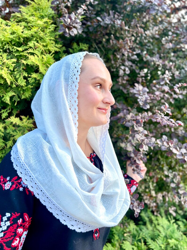 May include: A white headscarf with a delicate lace trim, draped around the neck. The scarf is paired with a black top featuring red and white embroidered floral patterns on the sleeves and neckline. The background includes green and purple foliage.