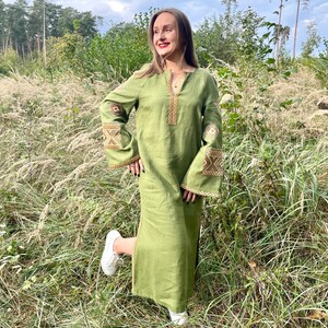 Pode incluir: Um vestido longo de linho verde-oliva com detalhes bordados nas mangas e decote. O vestido apresenta um corte folgado e mangas largas. A modelo est&aacute; usando t&ecirc;nis brancos. O vestido &eacute; adequado para uso casual.
