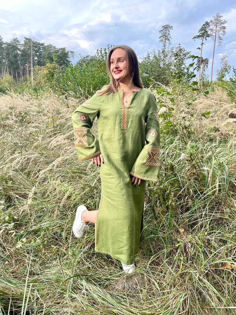 Pode incluir: Um vestido longo de linho verde-oliva com detalhes bordados nas mangas e decote. O vestido apresenta um corte folgado e mangas largas. A modelo est&aacute; usando t&ecirc;nis brancos. O vestido &eacute; adequado para uso casual.