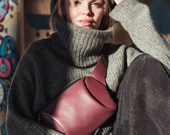 May include: A burgundy leather bum bag with a black zip and flap closure. The bag is worn over a grey turtleneck sweater. The person is wearing dark brown corduroy trousers.