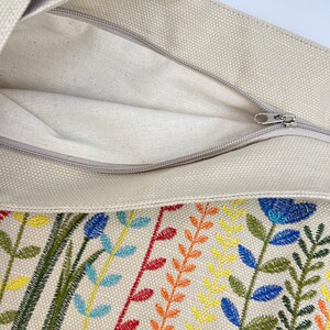 Puede incluir: Una bolsa de lona beige con cierre de cremallera. La bolsa presenta un dise&ntilde;o floral bordado de colores con hilo verde, azul, amarillo, naranja y rojo.