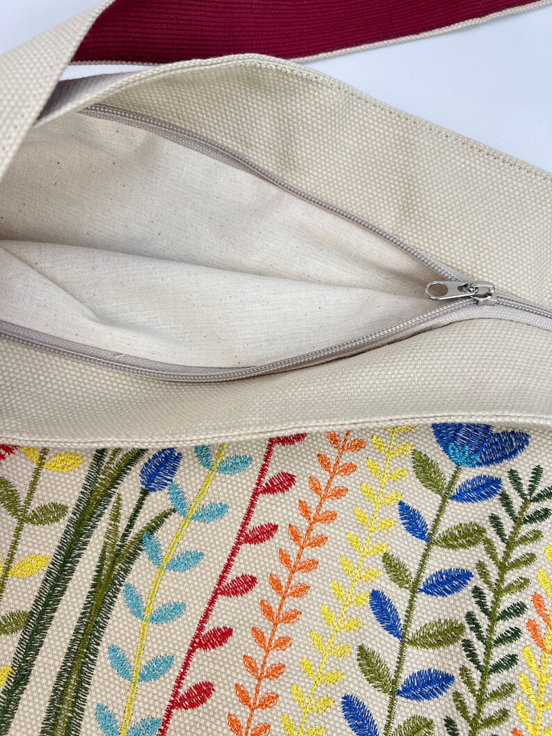 Puede incluir: Una bolsa de lona beige con cierre de cremallera. La bolsa presenta un dise&ntilde;o floral bordado de colores con hilo verde, azul, amarillo, naranja y rojo.