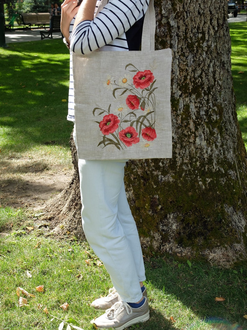 Op de afbeelding: Een linnen tas met geborduurde rode klaprozen en witte madeliefjes. De tas heeft een natuurlijke linnen kleur en wordt vastgehouden door iemand die een gestreept shirt en een witte broek draagt.