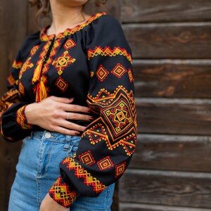 May include: A black embroidered blouse with red and yellow geometric patterns. The blouse has a long sleeve and a round neckline. The model is wearing a denim skirt.