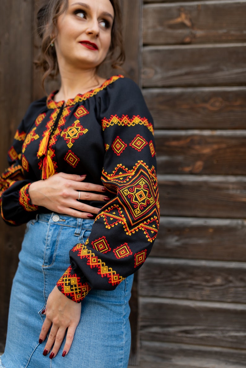 May include: A black embroidered blouse with red and yellow geometric patterns. The blouse has a long sleeve and a round neckline. The model is wearing a denim skirt.